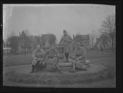 Militaires : quatre hommes en habit militaire autour d'une fontaine en pierre dans un parc ; maisons et enfants en arrière-plan