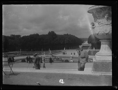 Versailles (Yvelines) : vue d'ensemble du bassin de Latone au centre du jardin du château de Versailles ; au premier plan deux hommes en habit militaire sont debout près du vase du soleil placé à l'extrémité occidentale du Parterre d'eau ; des visiteurs se promènent près du bassin